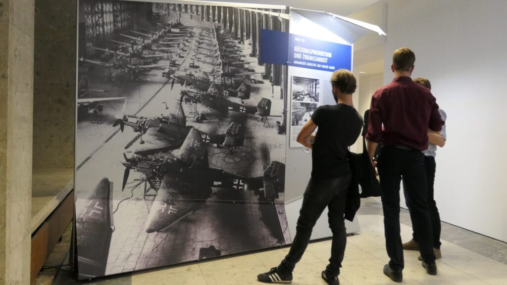 Exhibition visitors stand in front of an information panel about arms production.
